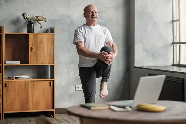 An older man stands in front of a laptop as he raises/stretches his knee to his chest.