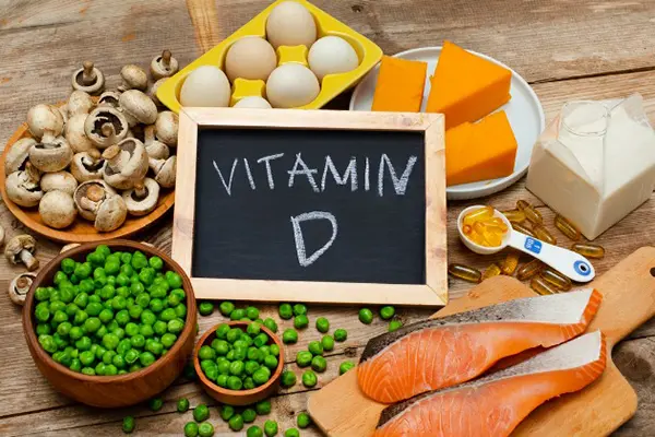 Vitamin D-rich foods on table surround a small chalkboard that reads Vitamin D. Foods include, salmon, a carton of milk, peas, mushrooms, eggs, cheese, and a tablespoon of vitamin d pill supplements. 