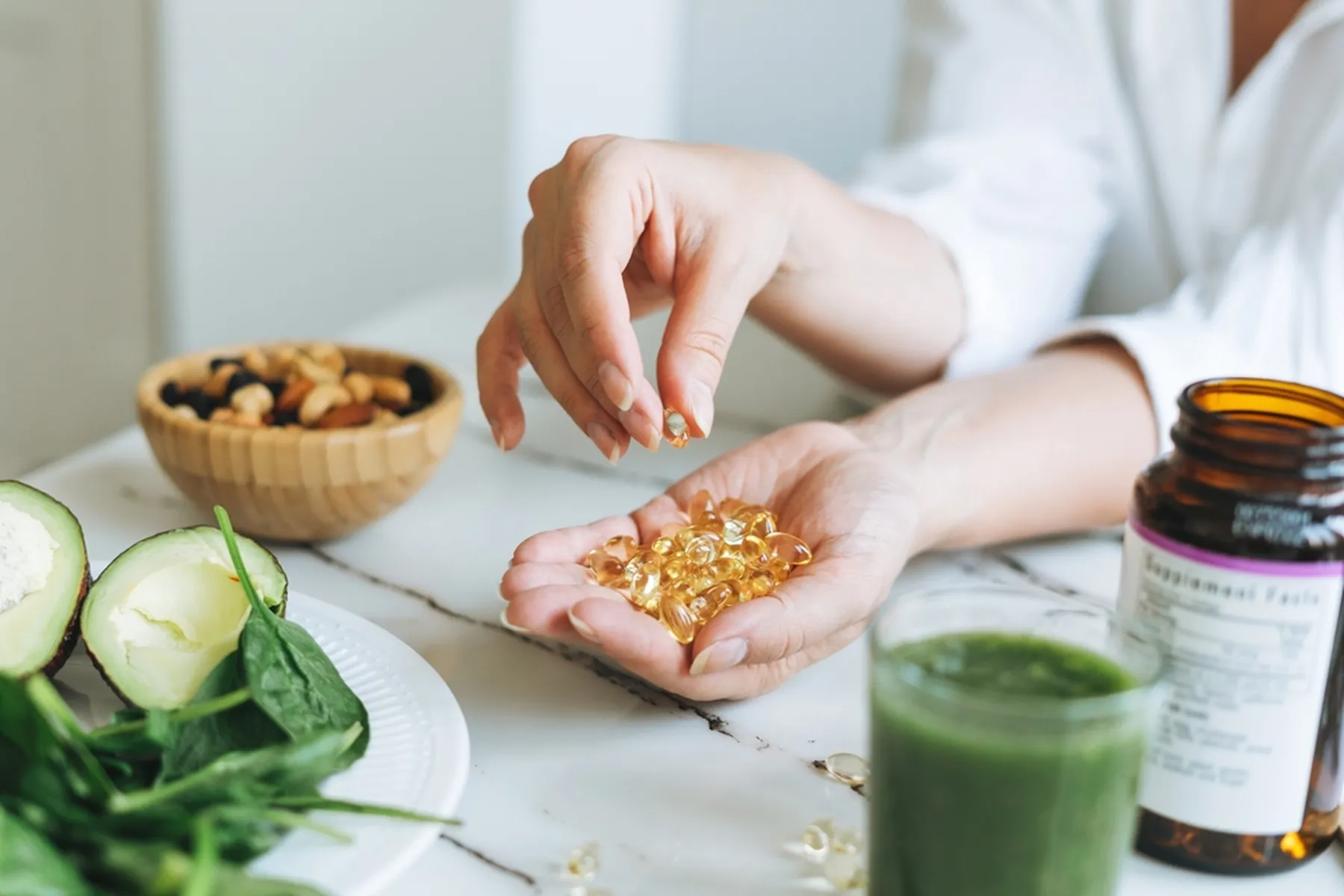 A woman holds vitamin d supplements in her hand, with a bowl of nuts and a green smoothie nearby.