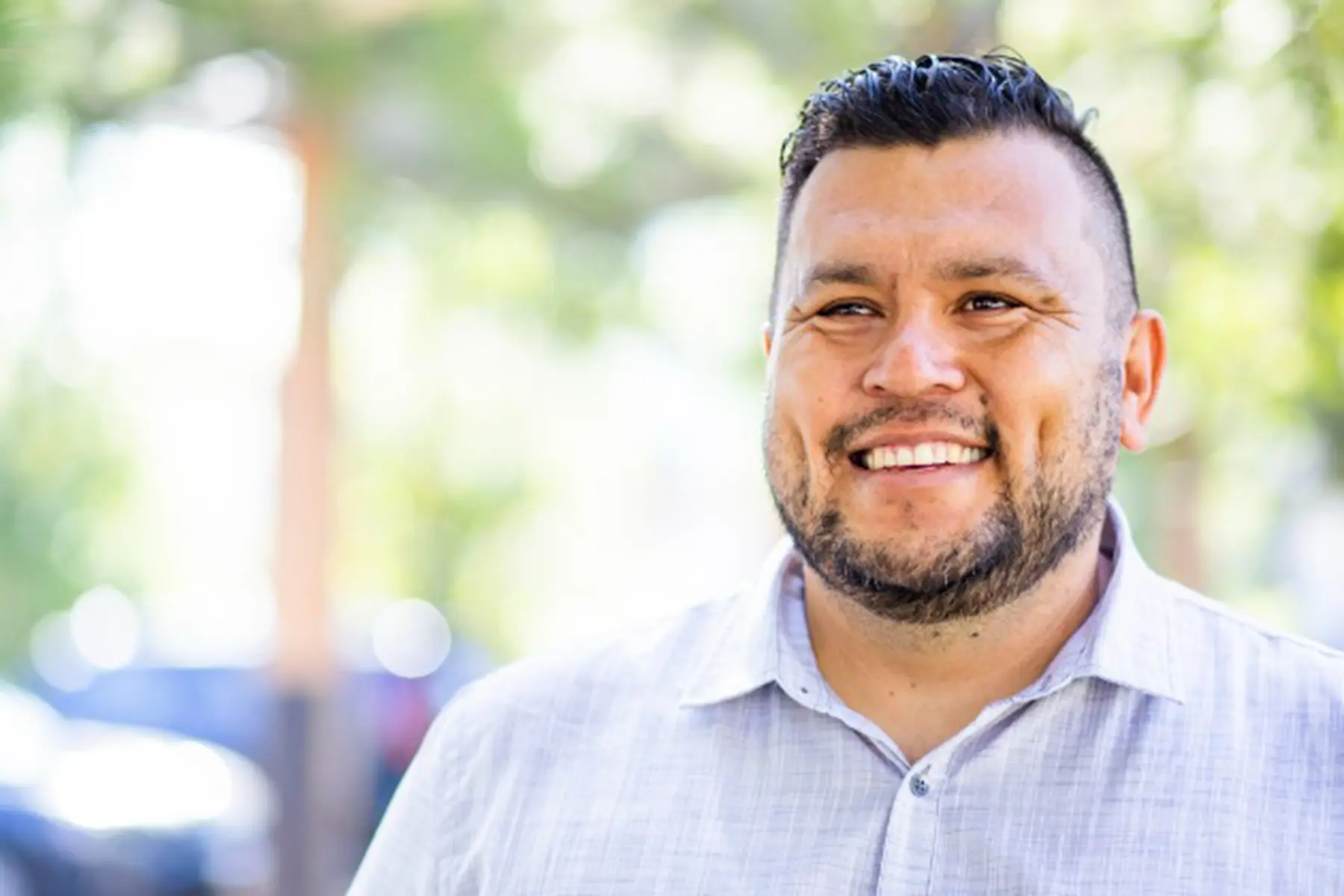 A young man in a button-down shirt smiles while outside. 