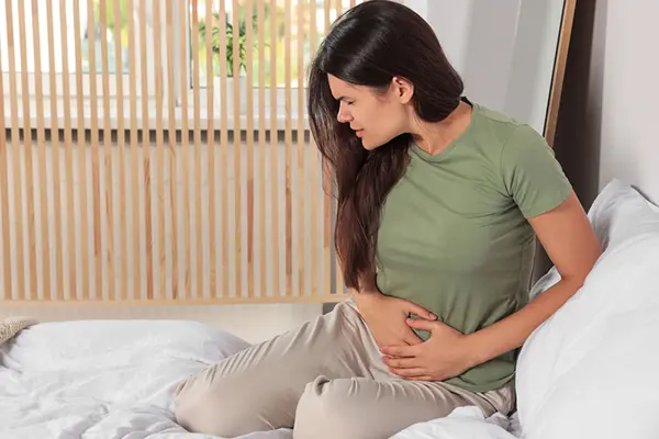 Woman sits on her bed and clutches her pelvis in pain.