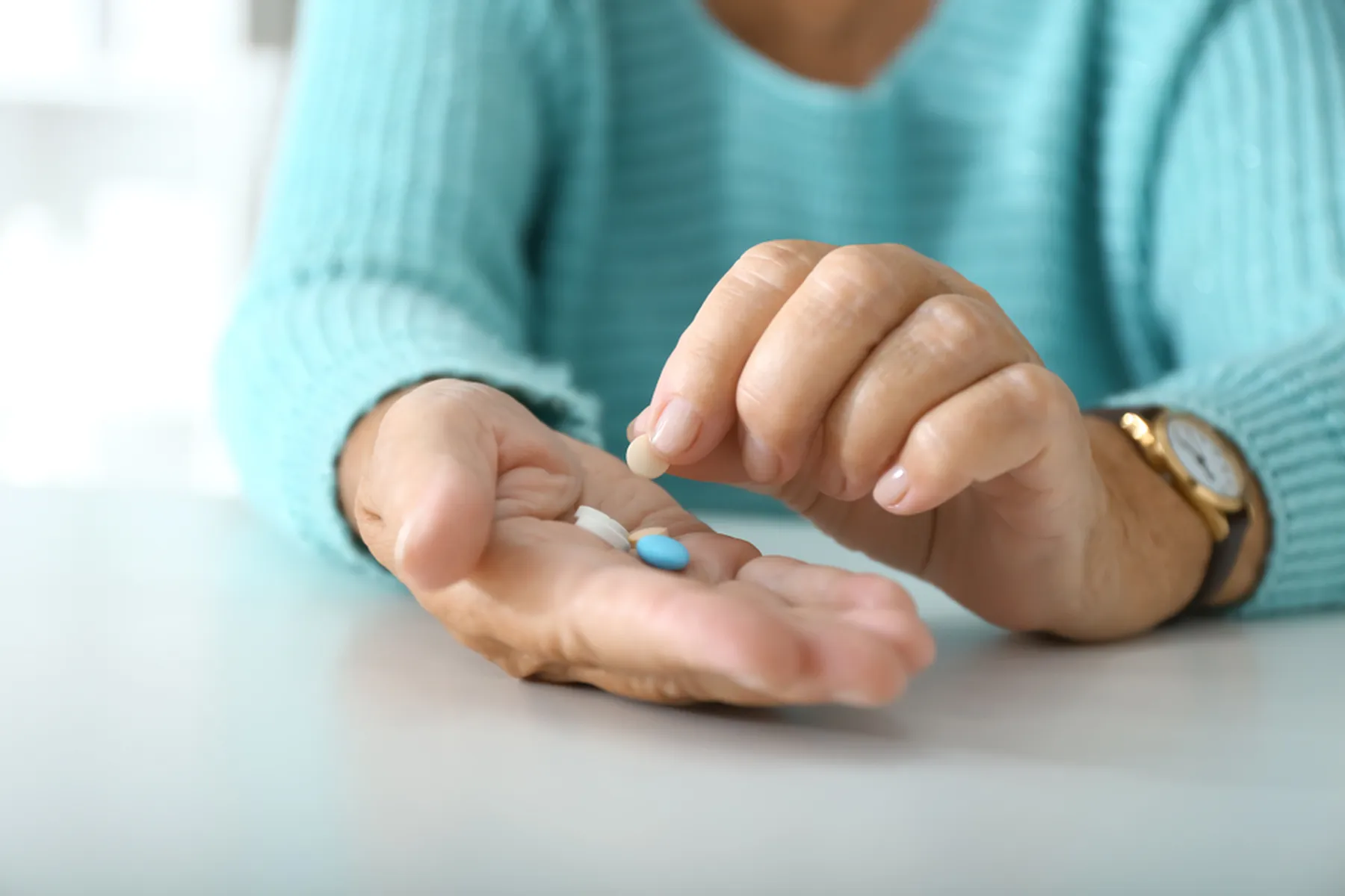 A woman in a light aqua sweater holding a few pills in her hand. She has a watch on her left wrist. 