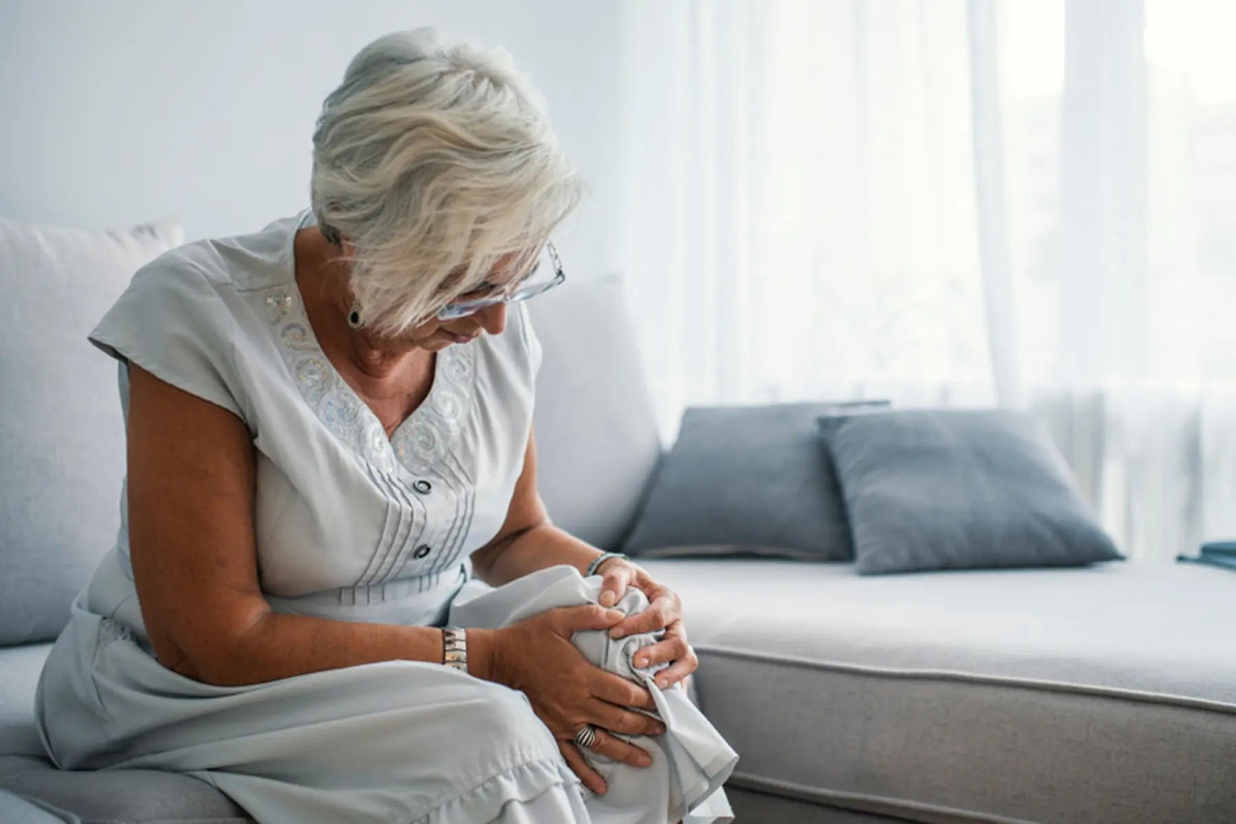 Middle aged woman sitting on couch and gripping her knee in pain.