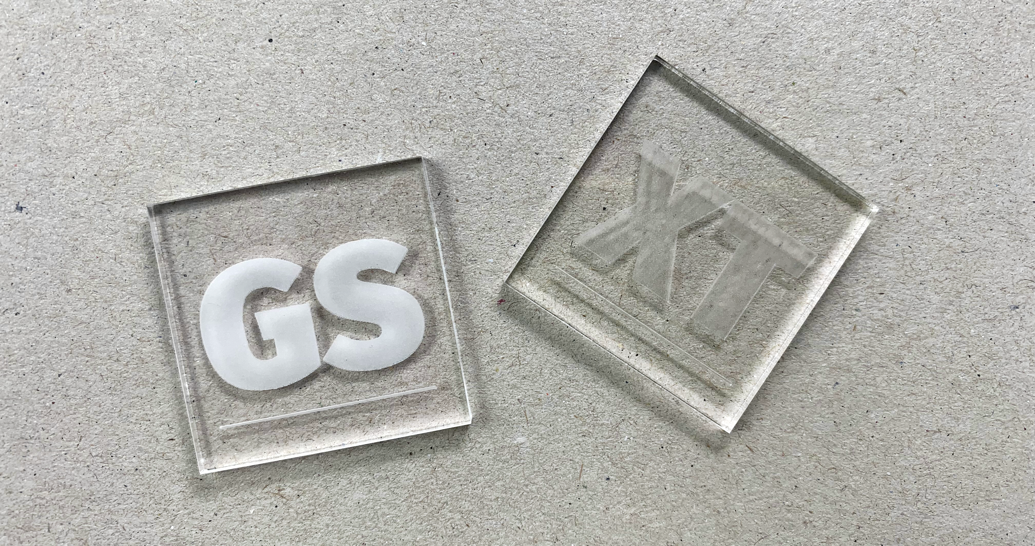 Two small laser-cut squares (approximately 2x2cm) of transparent acrylic. The left one is made of cast (GS) acrylic and the right one is made of extruded (XT) acrylic. The letters GS and XT are laser engraved on both. The GS engraving is matte white and high contrast, the XT engraving is less contrasty. This shows the difference in engraving quality between these two differently produced materials.