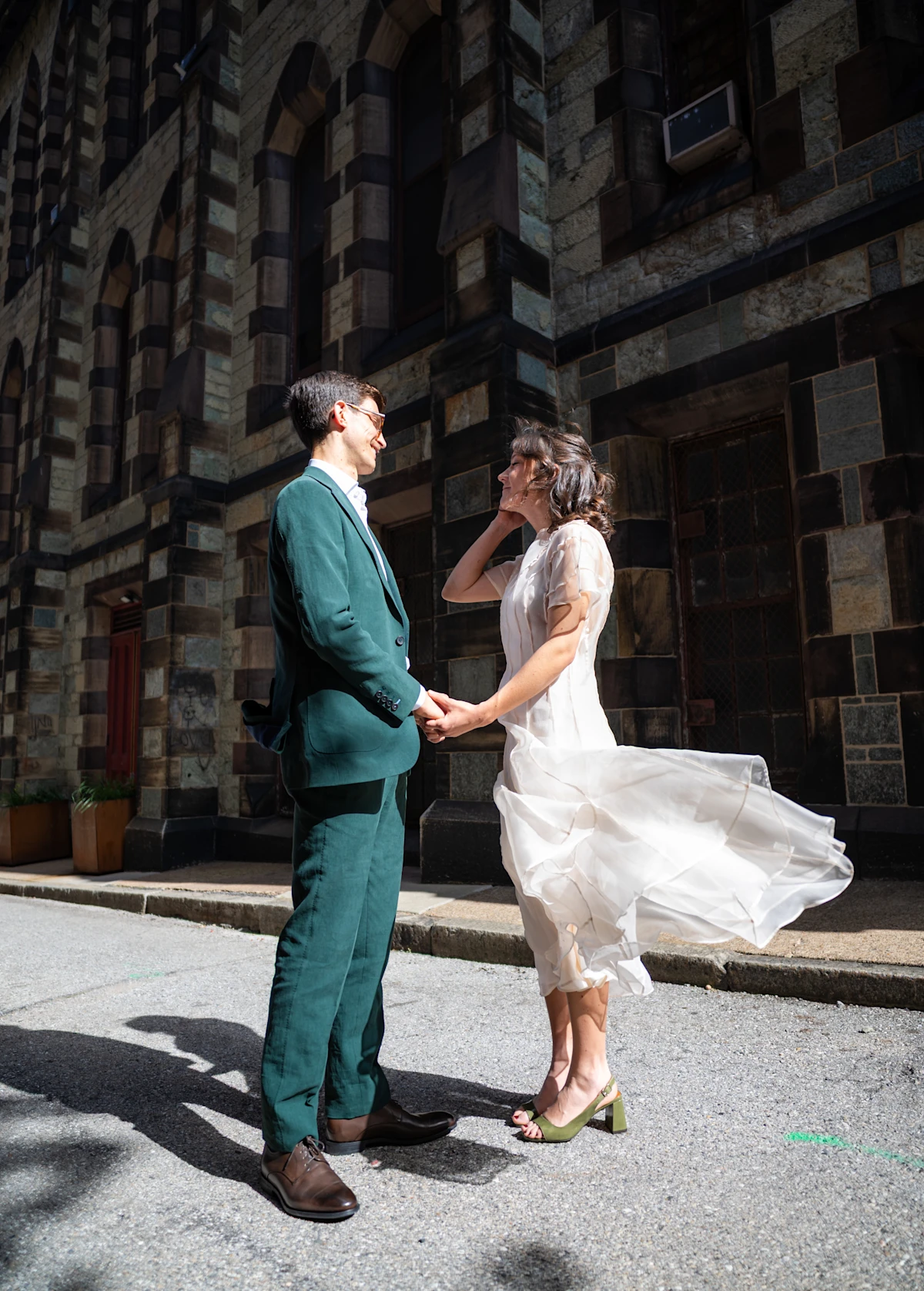A groom in green holds hands with a bride in a flowing white dress in the wind.