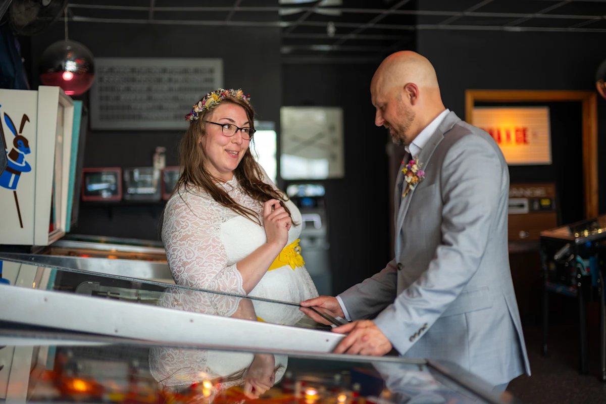 Bride teasing groom while he plays pinball.