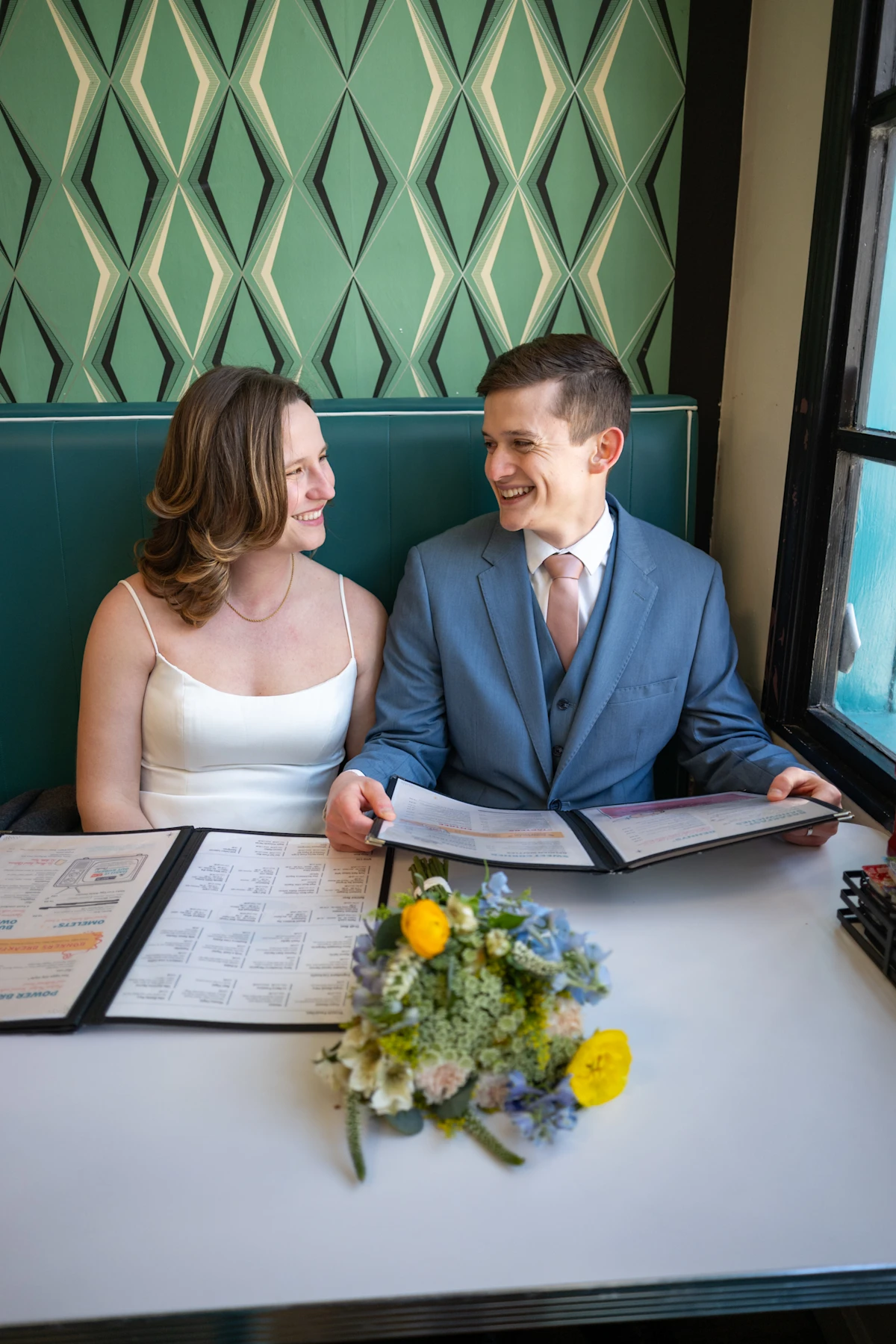 Bride and groom smile at each other as they sit at a table in a diner.