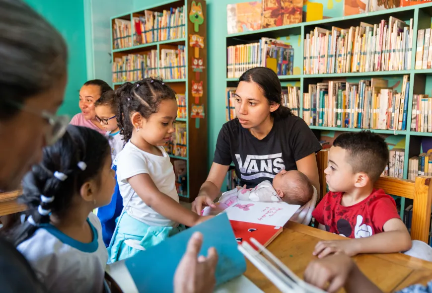 Cuatro niños y dos adultos se reúnen alrededor de una mesa que tiene libros. Están en una biblioteca.