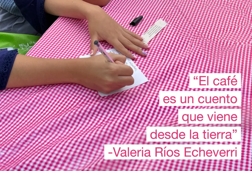 Un par de manos. Una apoyada sobre una mesa con mantel de cuadros, sosteniendo un papel, la otra escribiendo un verso. Una cita agregada sobre el lado derecho en un cuadro dice "El café es un cuento que viene desde la tierra" -Valeria Ríos Echeverri