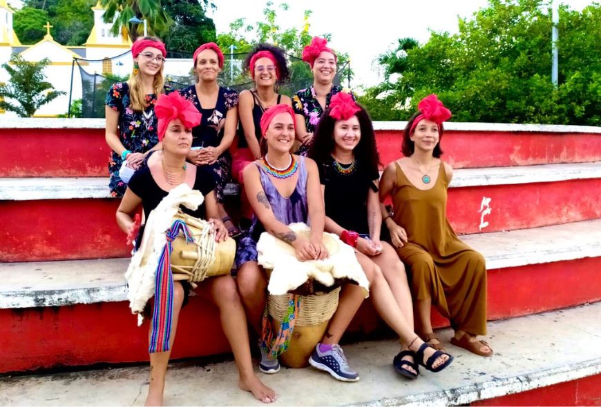 Ocho mujeres com tambores sentadas en unas gradas sonríen a la cámara.