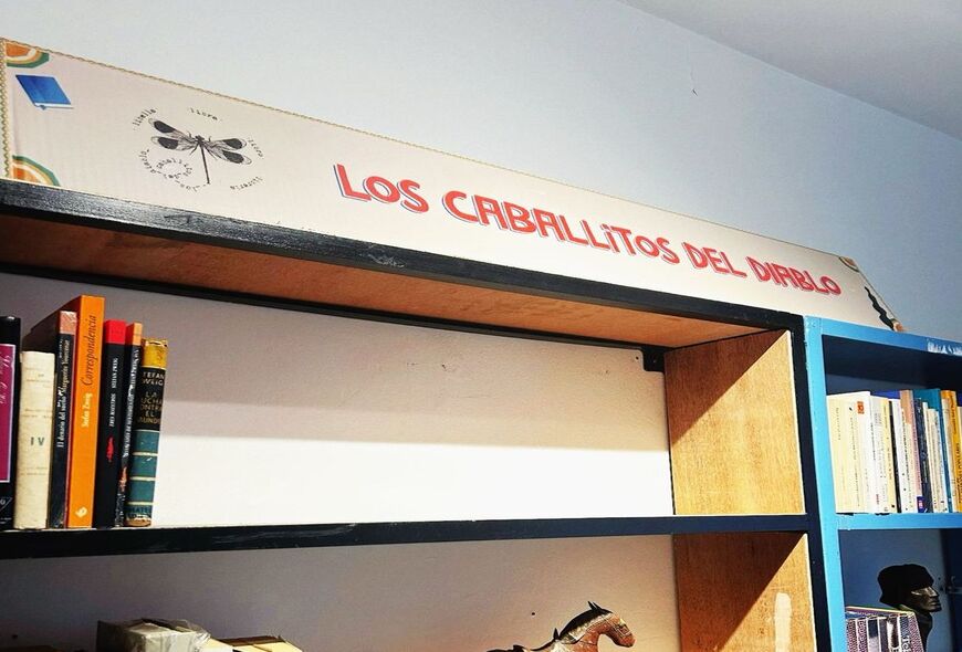 Cartel con el nombre de la librería Los Caballitos del Diablo, con el logo de la librería al lado izquierdo, una ilustración de una libélula con las palabras "liberal. libra. libro. librería".