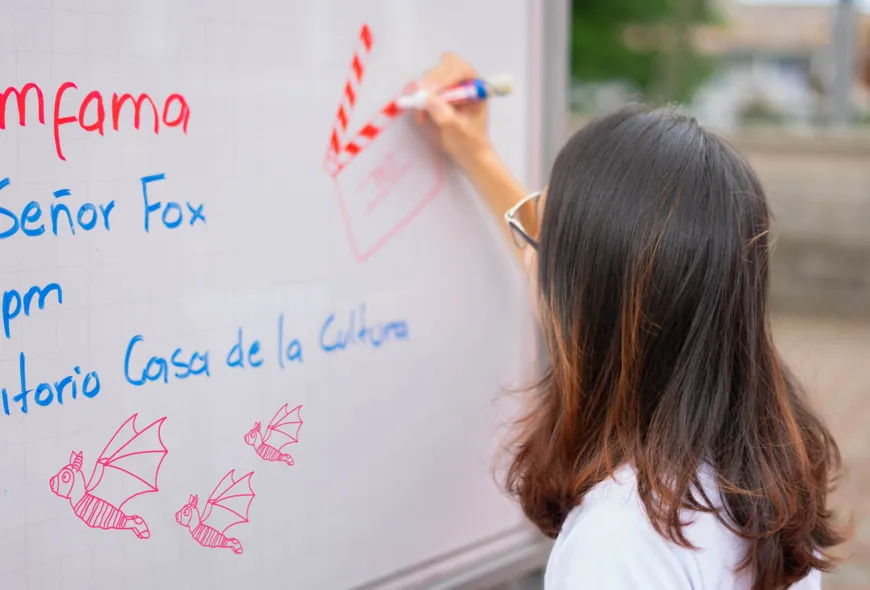 Una mujer con cabello oscuro hasta los hombros, rellena un dibujo de una claqueta en un tablero blanco. Se ve parte de la programación del evento, la invitación a la proyección de la película "El Increíble Señor Fox"