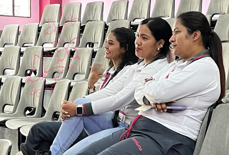 Tres mujeres sentadas en un auditorio miradas desde el lado. La más alejada de la cámara mueve la mano con emoción sobre su pecho. Las tres sonríen a la ponente, fuera de toma