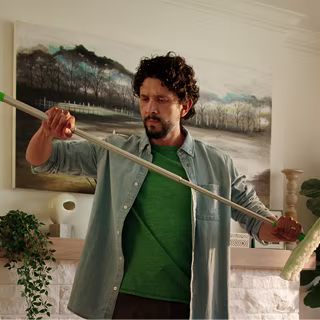 A man standing in a living room holding an extended paint roller pole, with home décor and plants visible in the background.