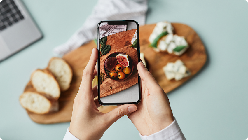 Hands holding a phone which is taking a photo of the center of a charcuterie board on the surface below it.