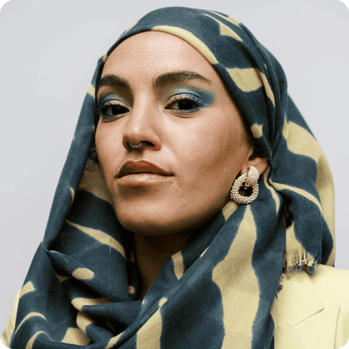 Woman wearing pearl encrusted earrings, a dark green and yellow print hijab with matching green eyeshadow and a nose ring