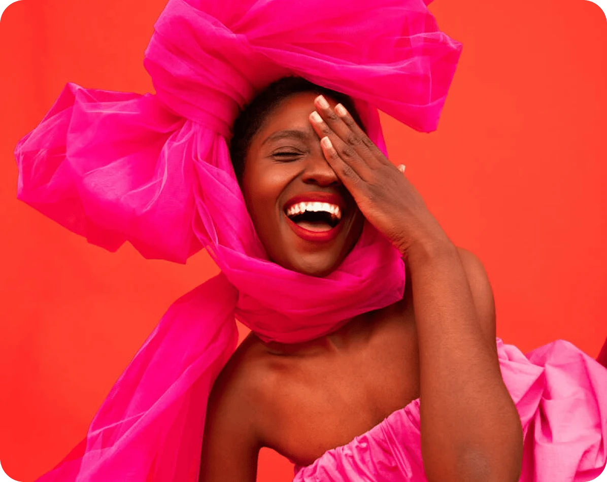 Woman smiling and covering one eye, wearing a pink dress and and an oversized pink bow on her head
