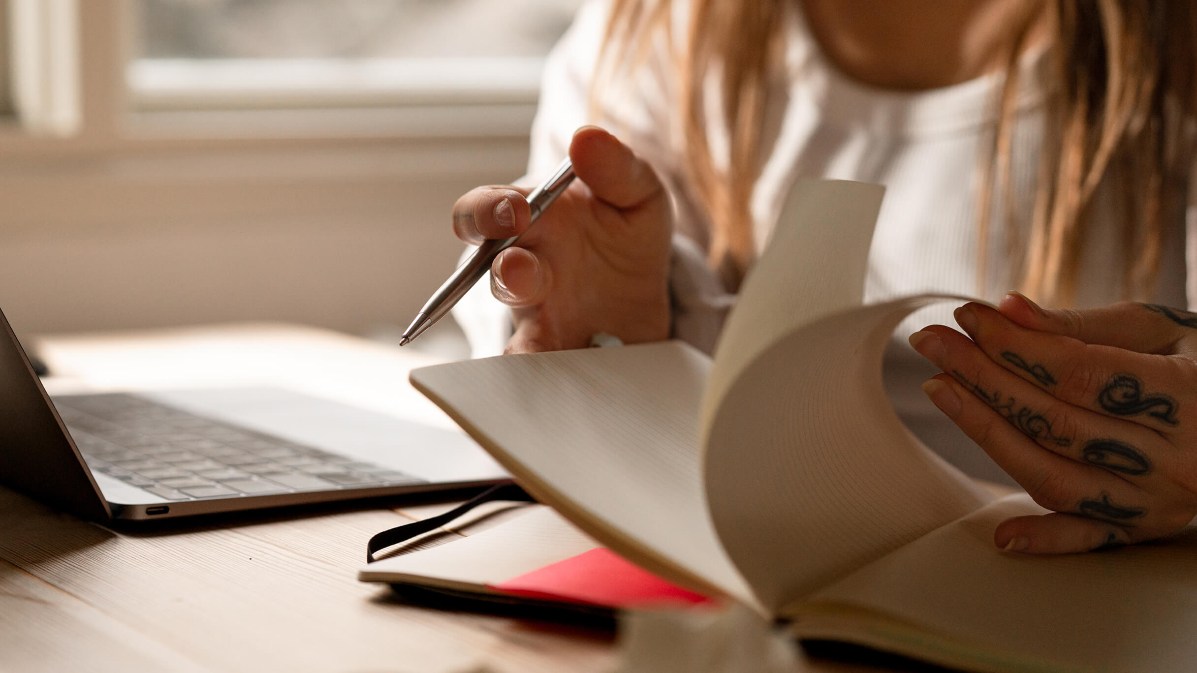 Close-up of a creator’s hands planning in a notebook beside a laptop at a warm, sunlit desk.