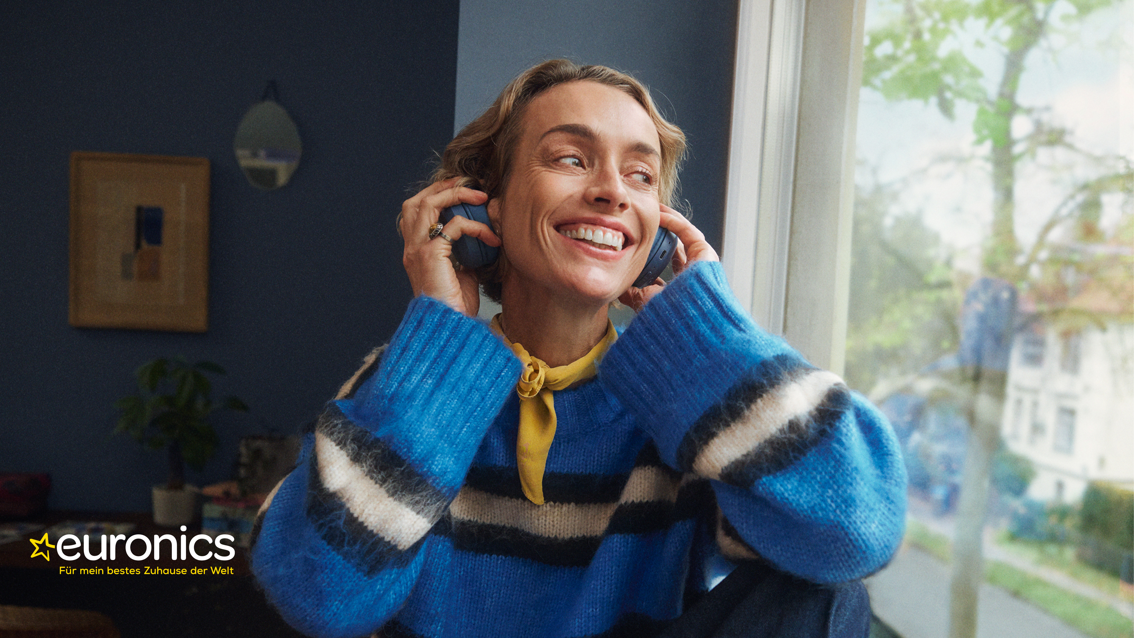 Person mit blau-weiß gestreiftem Pullover und gelbem Halstuch hält Kopfhörer an den Ohren, im Hintergrund Fenster