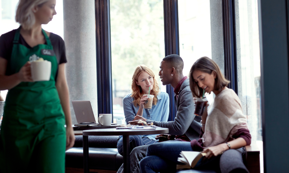Drei Personen sitzen in einem Café, trinken Kaffee und unterhalten sich, Laptop und Notizbuch auf dem Tisch.