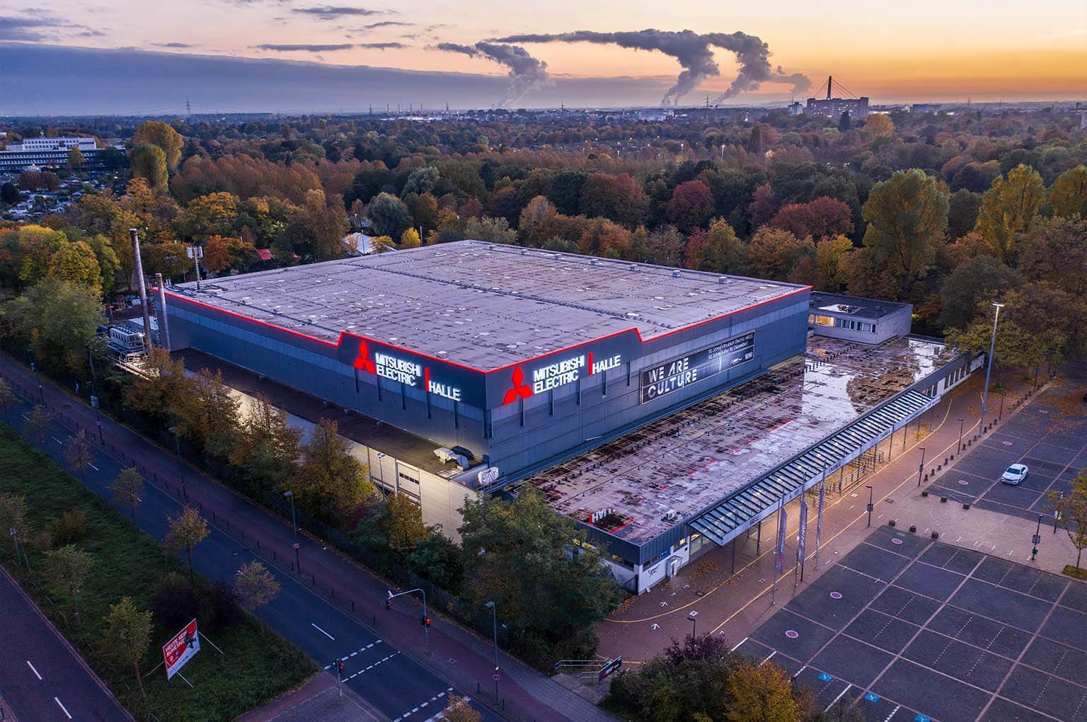 Mitsubishi Electric Halle umgeben von Bäumen, im Hintergrund Industrieanlagen mit rauchenden Schornsteinen