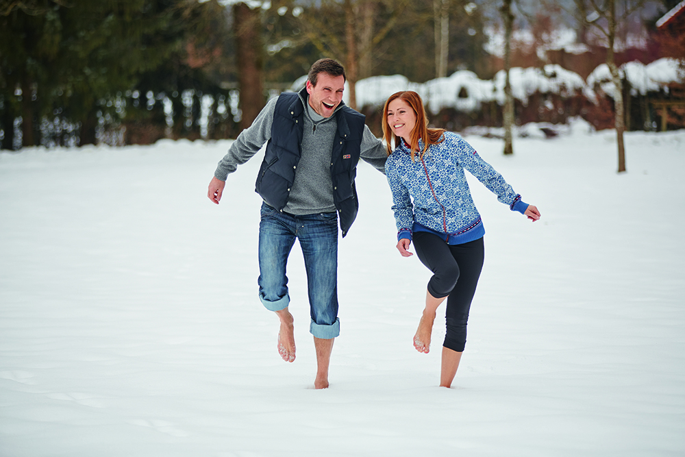 Zwei Personen laufen barfuß durch verschneite Landschaft, Bäume im Hintergrund