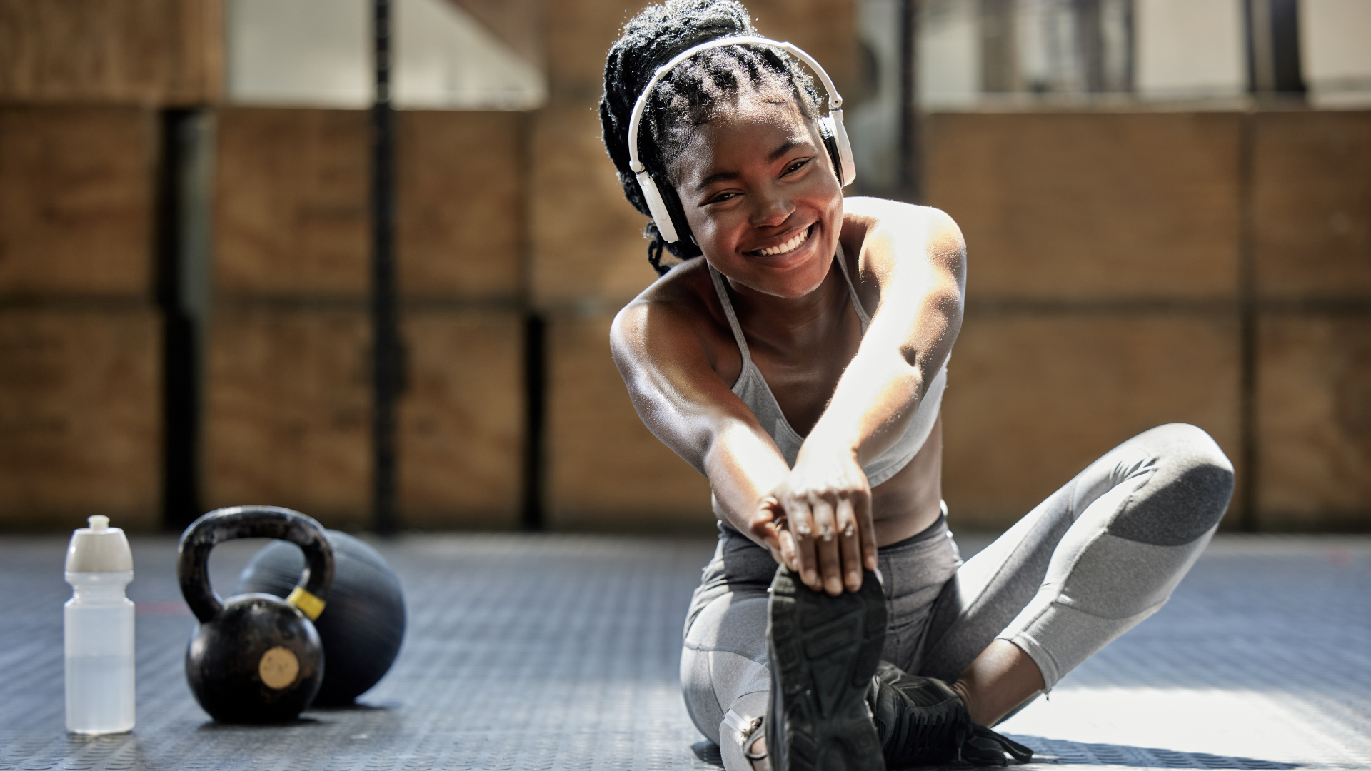 Frau mit Kopfhörern dehnt sich im Fitnessstudio, neben ihr Kettlebell und Wasserflasche
