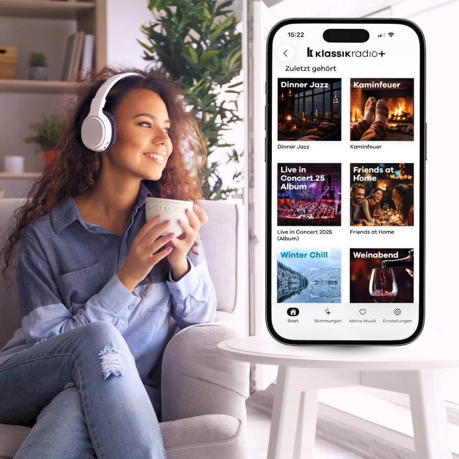 Smartphone mit geöffneter Klassik Radio Plus App liegt auf einem Notizbuch, daneben Kopfhörer, Stift und eine Tasse Kaffee.