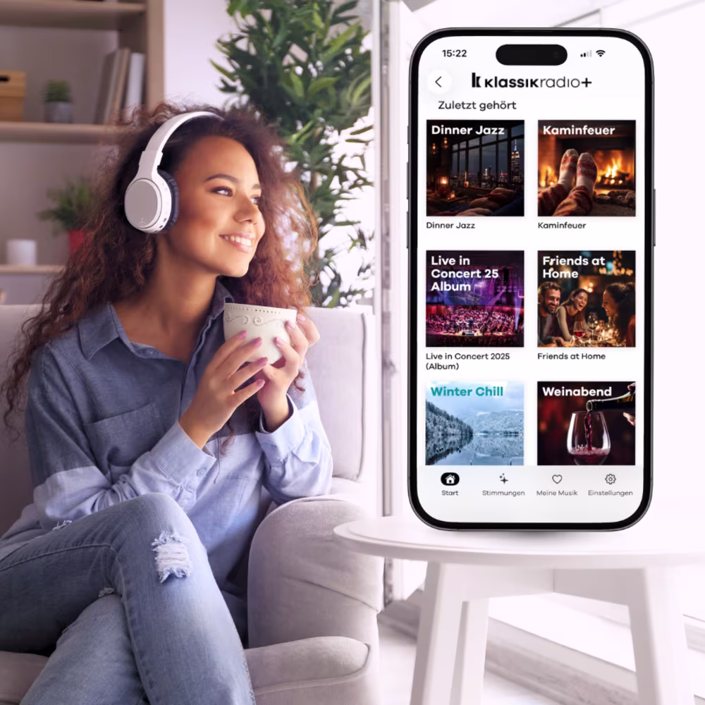 Smartphone mit geöffneter Klassik Radio Plus App liegt auf einem Notizbuch, daneben Kopfhörer, Stift und eine Tasse Kaffee.