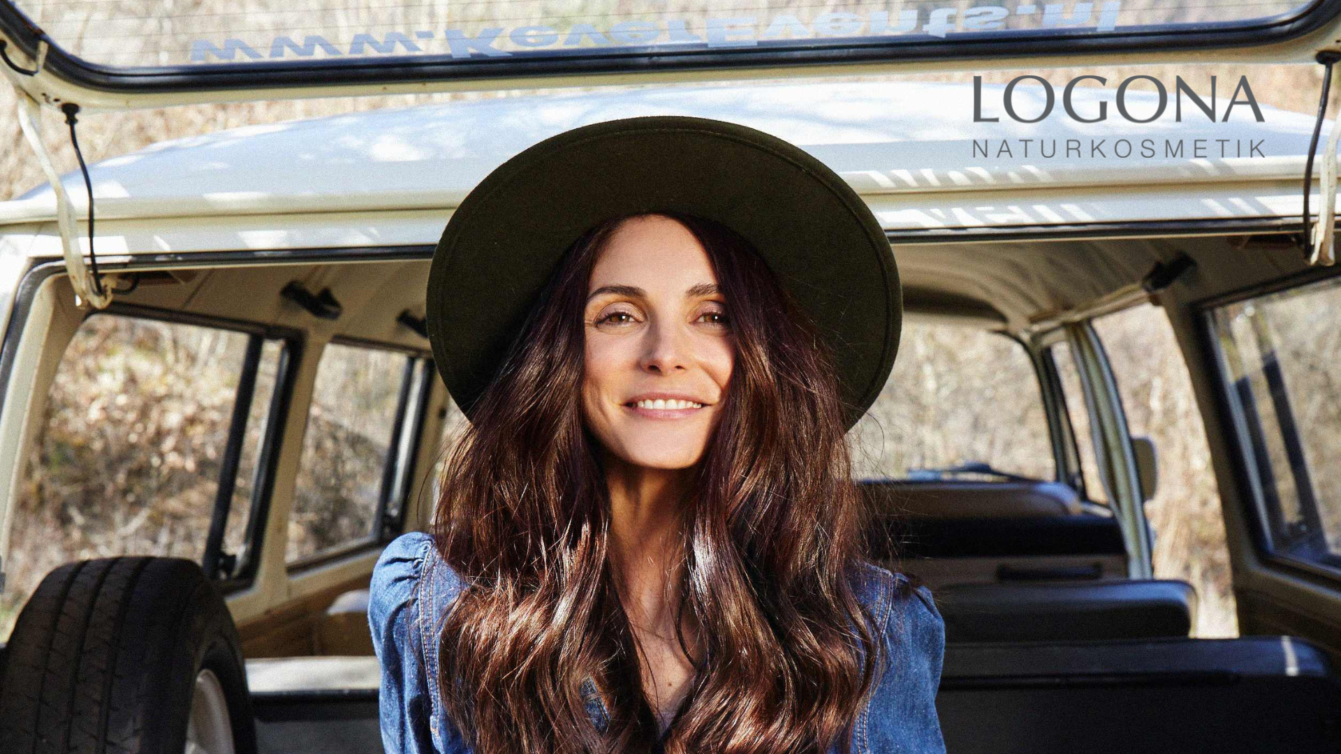 Person mit langen, gewellten braunen Haaren und grünem Hut sitzt in einem offenen Auto, LOGONA Naturkosmetik Schriftzug sichtbar