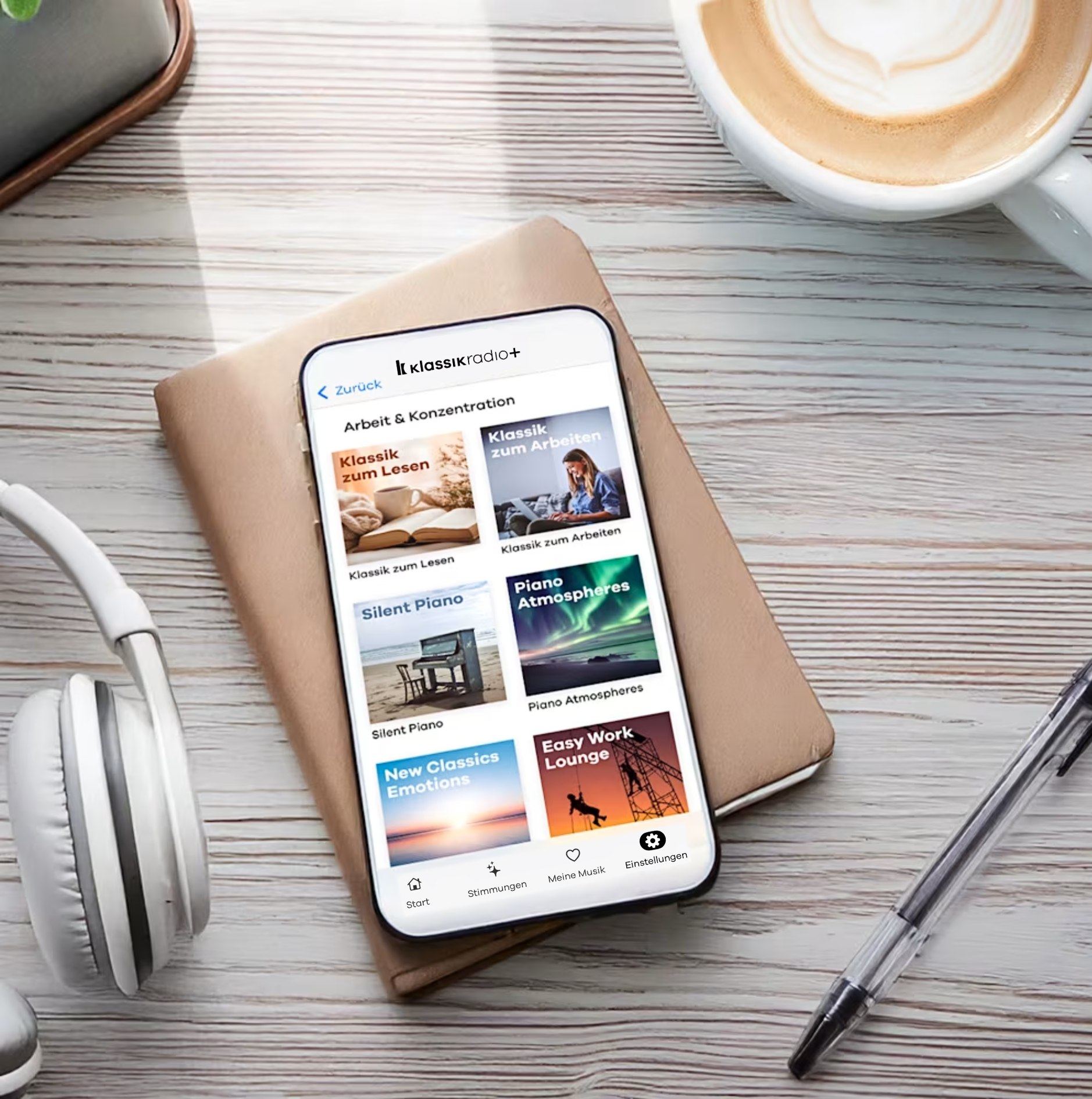 Smartphone mit der Klassik Radio Plus App geöffnet. Es liegt auf einem Schreibtisch mit Kaffee, Kopfhörern, Stift und Notizblock.