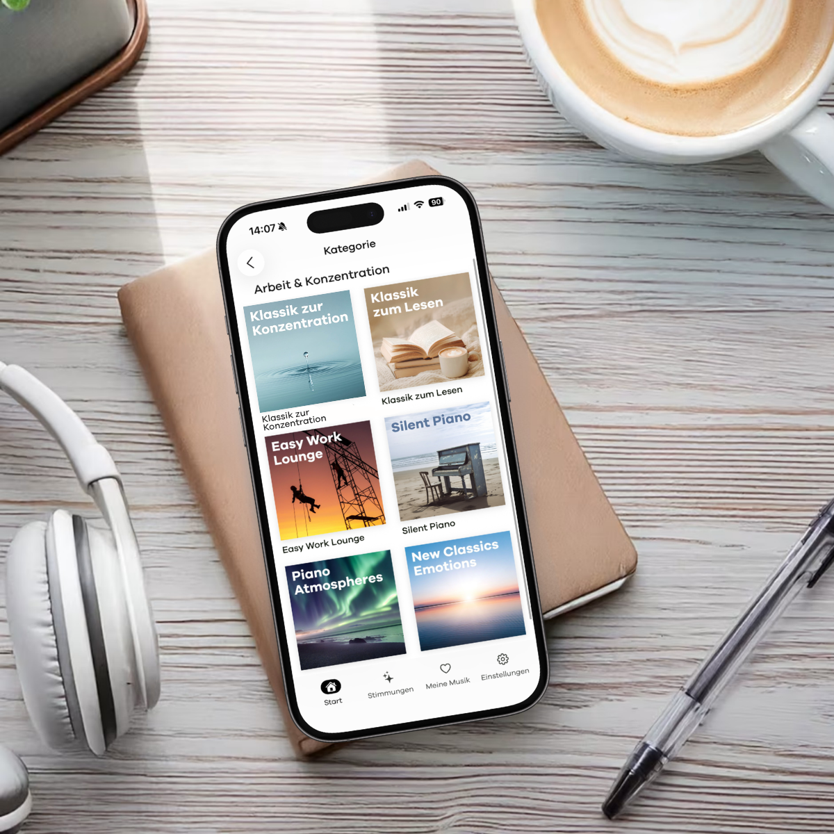 Smartphone mit der Klassik Radio Plus App geöffnet. Es liegt auf einem Schreibtisch mit Kaffee, Kopfhörern, Stift und Notizblock.