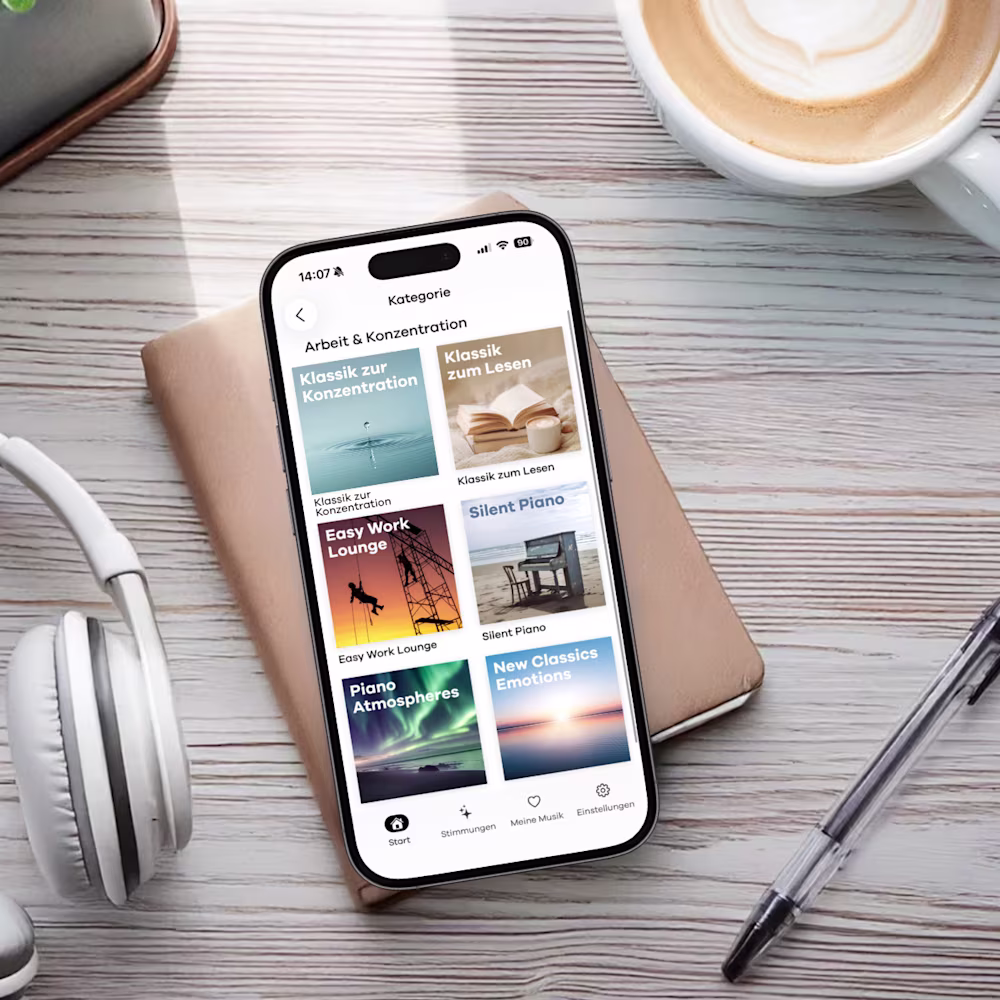 Smartphone mit der Klassik Radio Plus App geöffnet. Es liegt auf einem Schreibtisch mit Kaffee, Kopfhörern, Stift und Notizblock.