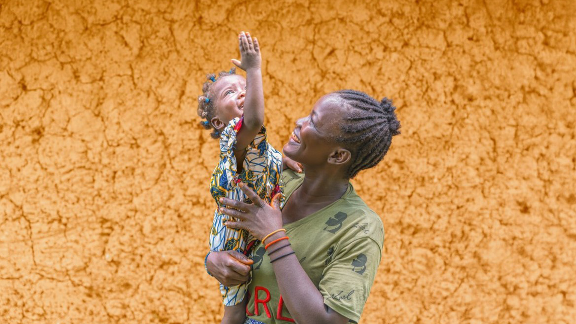 Frau hält ein Kleinkind vor einer Lehmwand, das Kind streckt den Arm nach oben aus