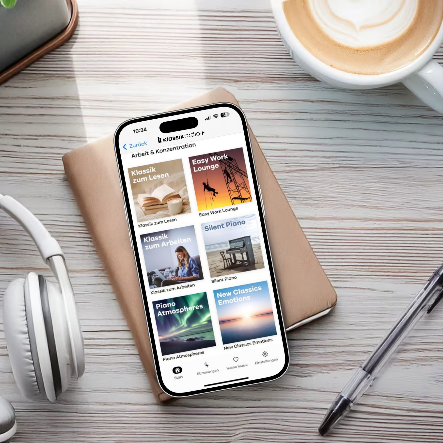 Smartphone mit geöffneter Klassik Radio Plus App liegt auf einem Notizbuch, daneben Kopfhörer, Stift und eine Tasse Kaffee.