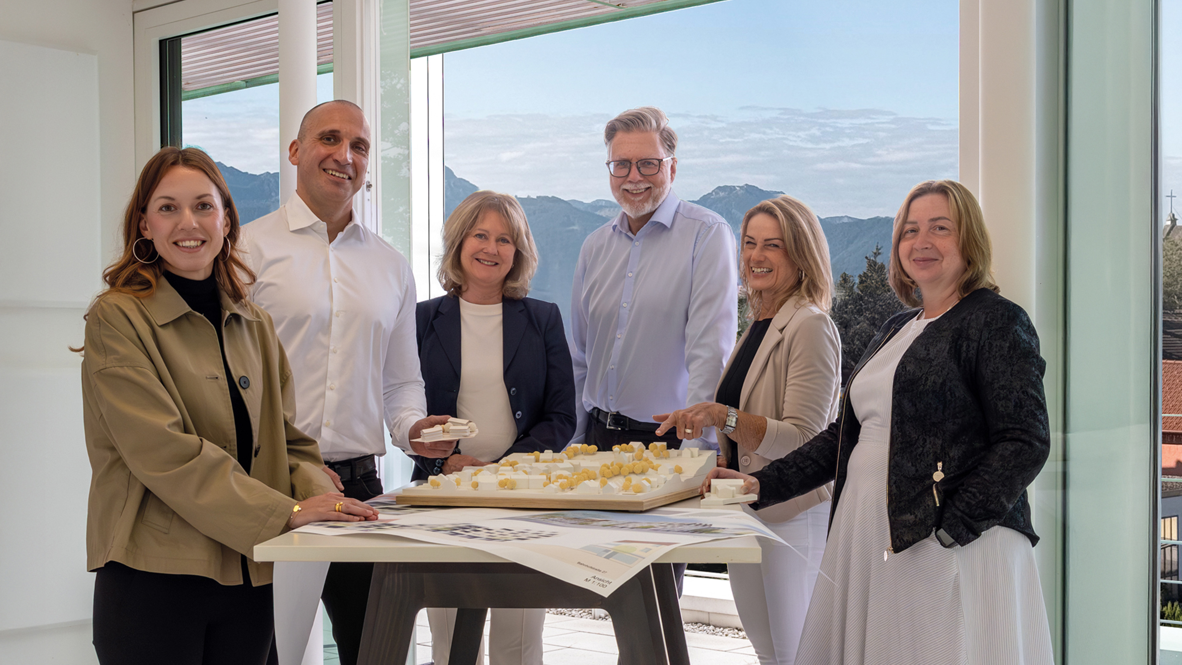 Sechs Personen stehen um einen Tisch mit Architekturmodell und Bauplänen in modernem Büro mit Bergblick.
