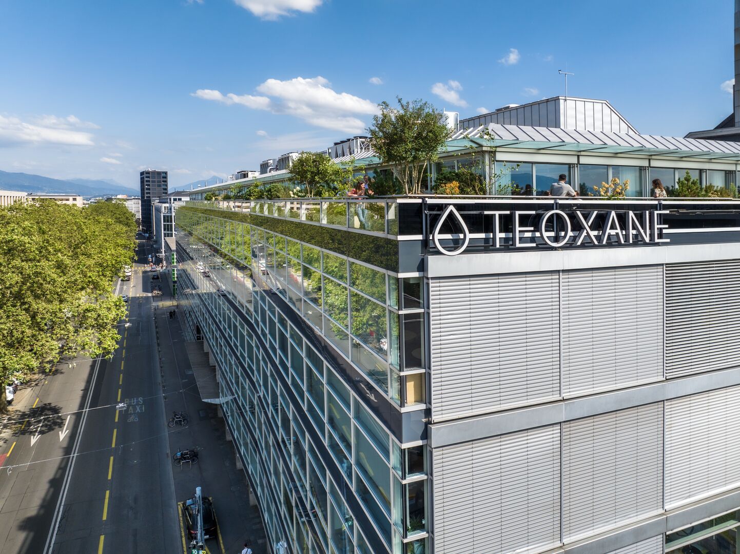Modernes Bürogebäude mit TEOXANE-Logo, begrünte Dachterrasse, Straße und Bäume im Hintergrund