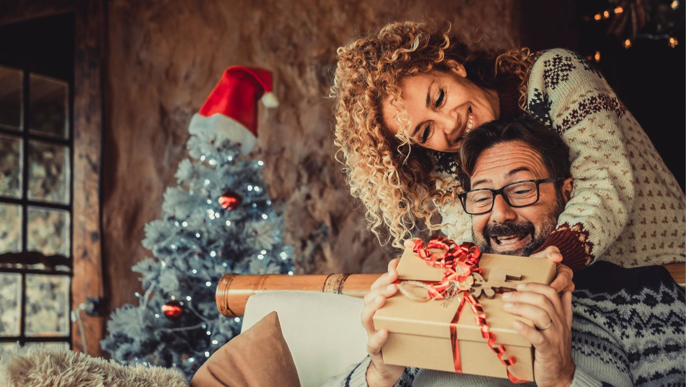 Mann wird neben dem Weihnachtsbaum von seiner Frau mit einem Geschenk überrascht