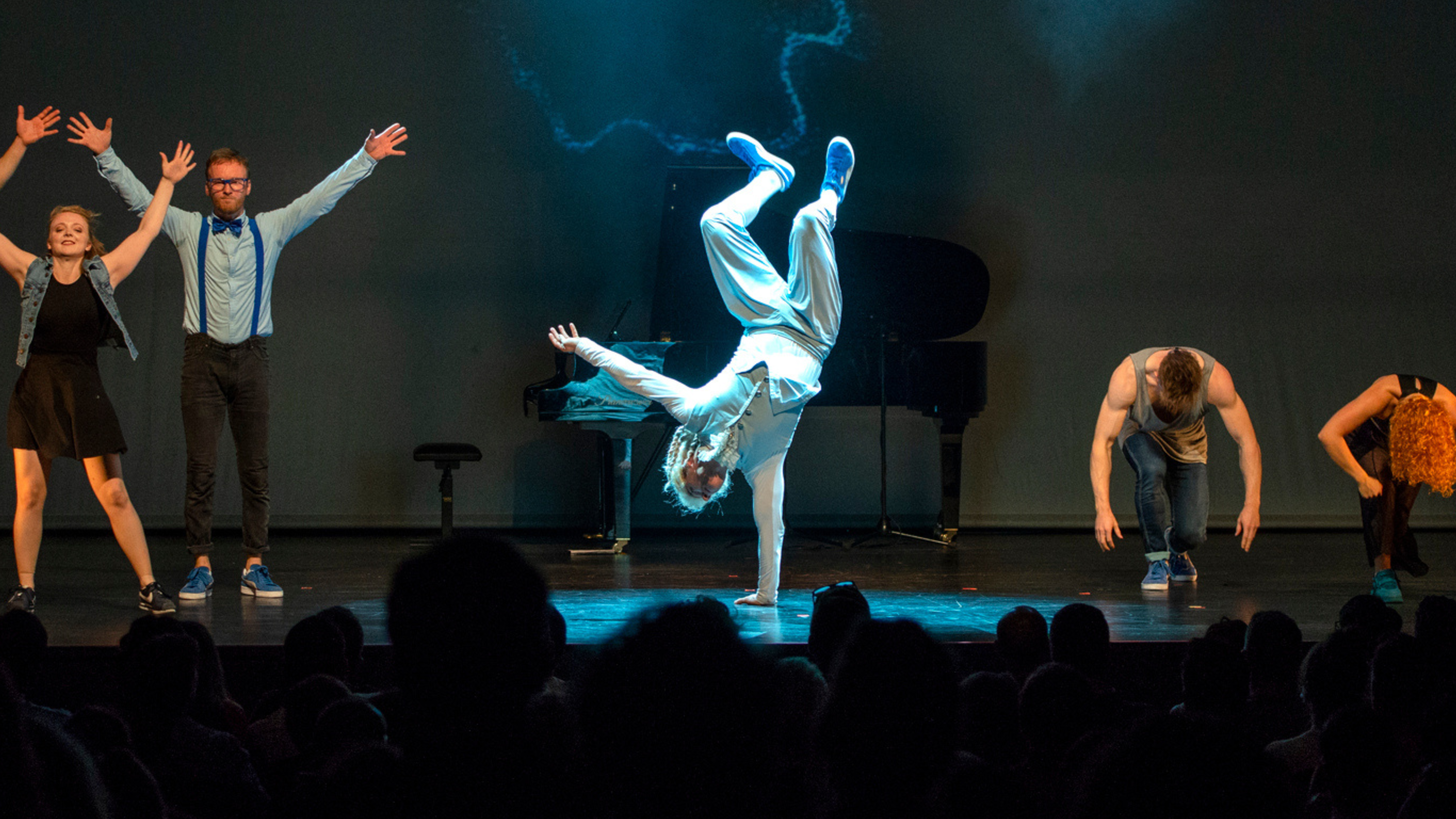 Tanzgruppe auf Bühne, eine Person macht einen Handstand, im Hintergrund ein Klavier