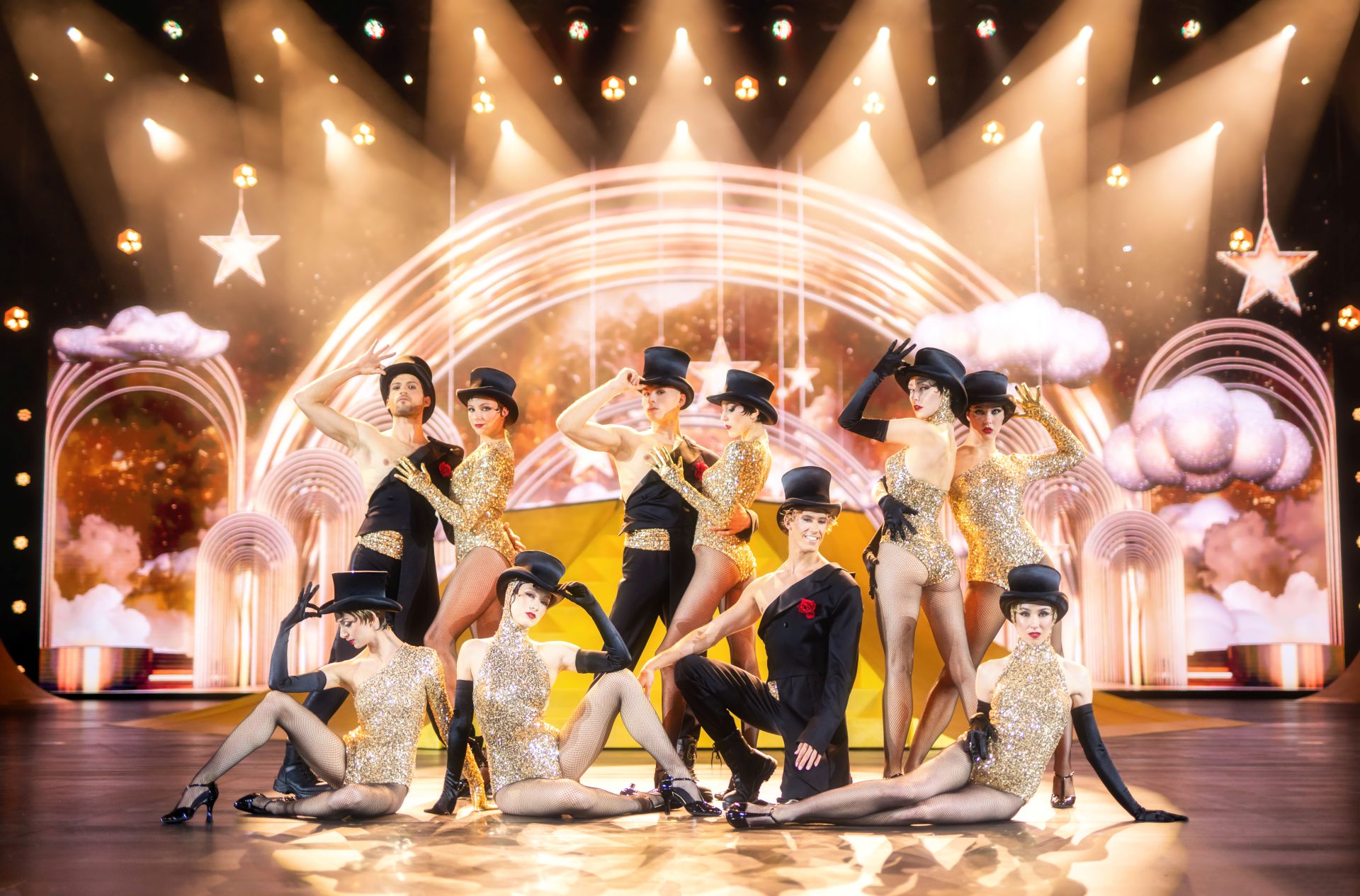 Tänzerinnen und Tänzer in goldenen Kostümen und Zylindern auf einer glamourösen Showbühne mit Lichtern und Sternen
