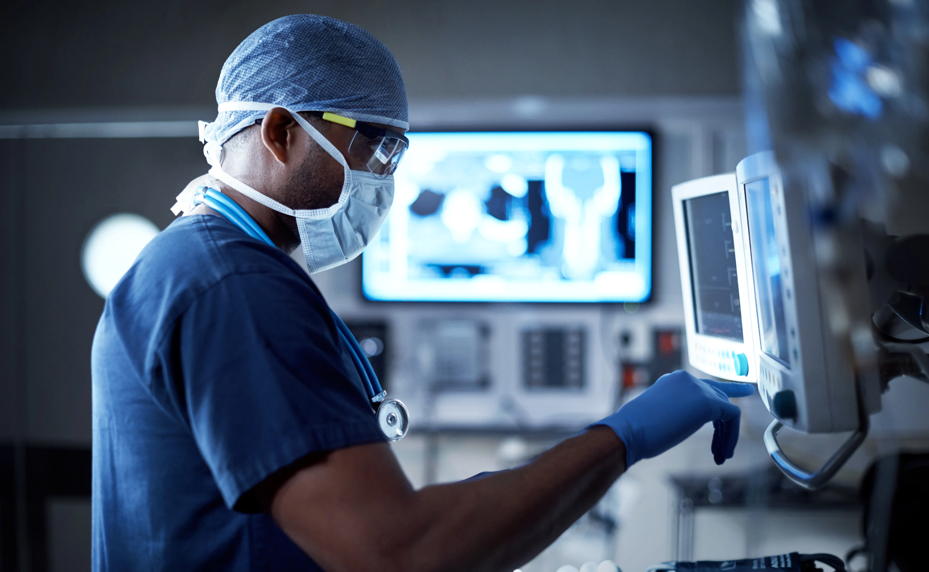 A healthcare employee in a mask and scrubs looks at a technical readout on a nearby monitor