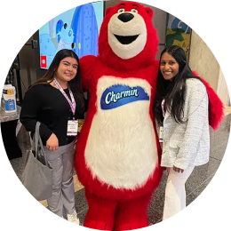 Deux femmes aux longs cheveux bruns, habillées en tenue de travail décontractée, posent avec une mascotte d'ours rouge de taille humaine pour la marque Charmin de P&G.