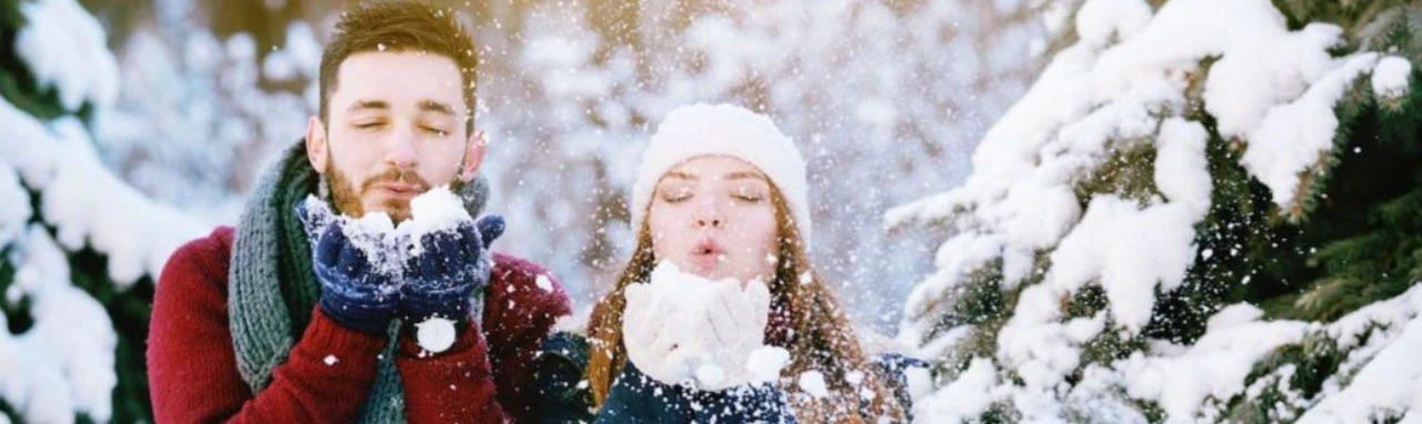Un couple se tient dans une forêt enneigée et souffle sur un petit tas de neige dans leurs mains