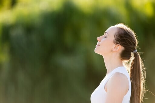 Une femme qui porte un débardeur, elle est brune et semble apaisée. Elle prend une grande respiration. Le fond est en majorité vert 