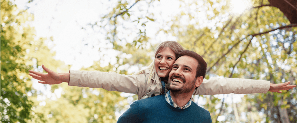 un homme porte une femme sur son dos, ils sourient. Des arbres en fond