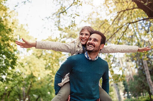 un homme porte une femme sur son dos, ils sourient. Des arbres en fond