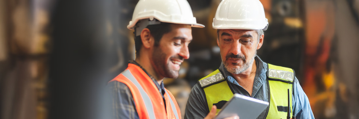 Two workers in hard hats and safety vests smiling while looking at a tablet in an industrial setting.