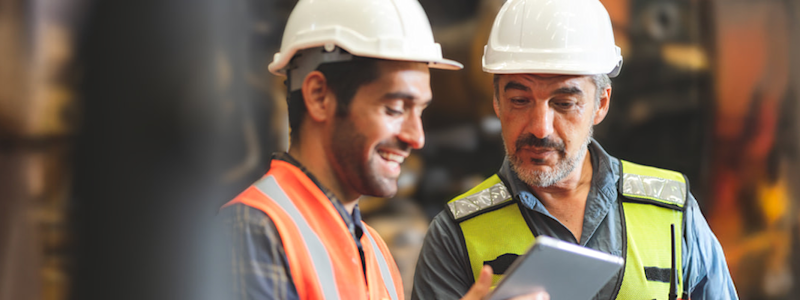 Two workers in hard hats and safety vests smiling while looking at a tablet in an industrial setting.