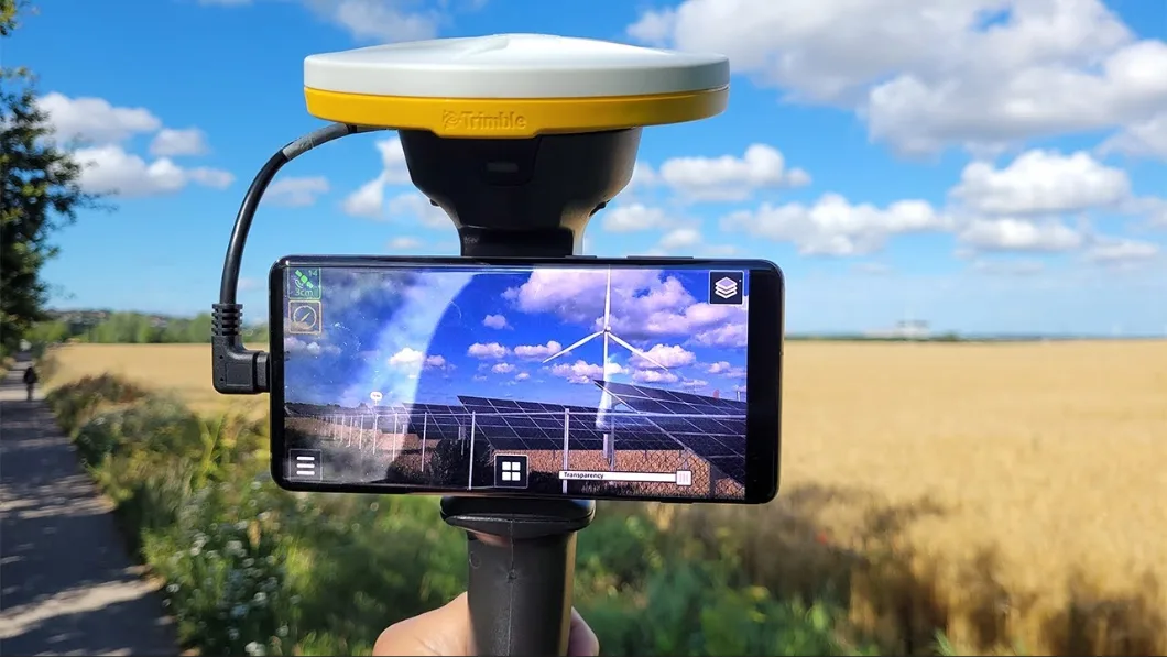 A Trimble GPS antenna mounted on a pole with a smartphone displaying solar panels and wind turbines in an AR surveying app.
