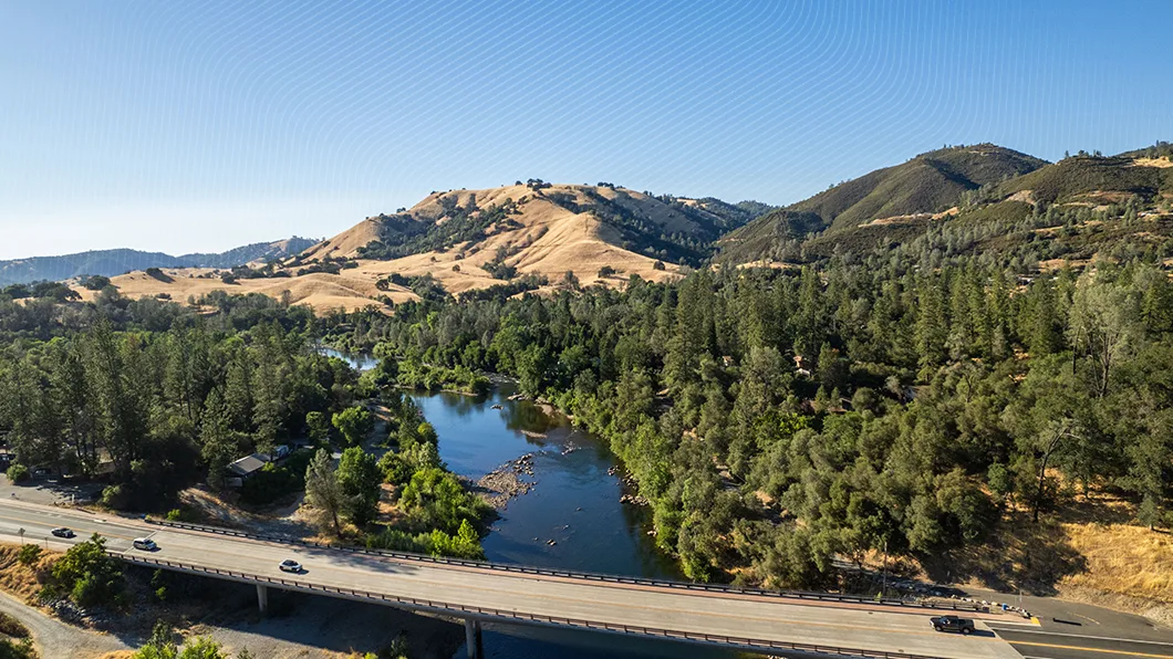 Aerial view of a multi-lane concrete highway bridge carrying vehicles over a river, surrounded by golden, tree-covered hills under a clear sky.